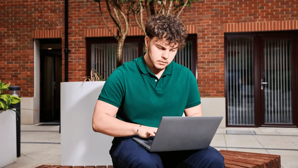 A young professional in a green polo shirt working on a laptop outdoors, representing a new hire engaging with their remote employee onboarding plan from a flexible workspace.