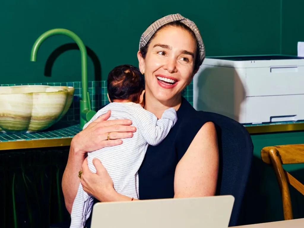 A professional woman smiling while working on a laptop and holding a baby, symbolizing the work-life balance enabled by a flexible remote work policy.