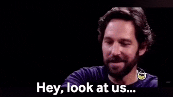 Man with a beard looking surprised and saying, 'Hey, look at us,' with a smile, expressing disbelief or amazement