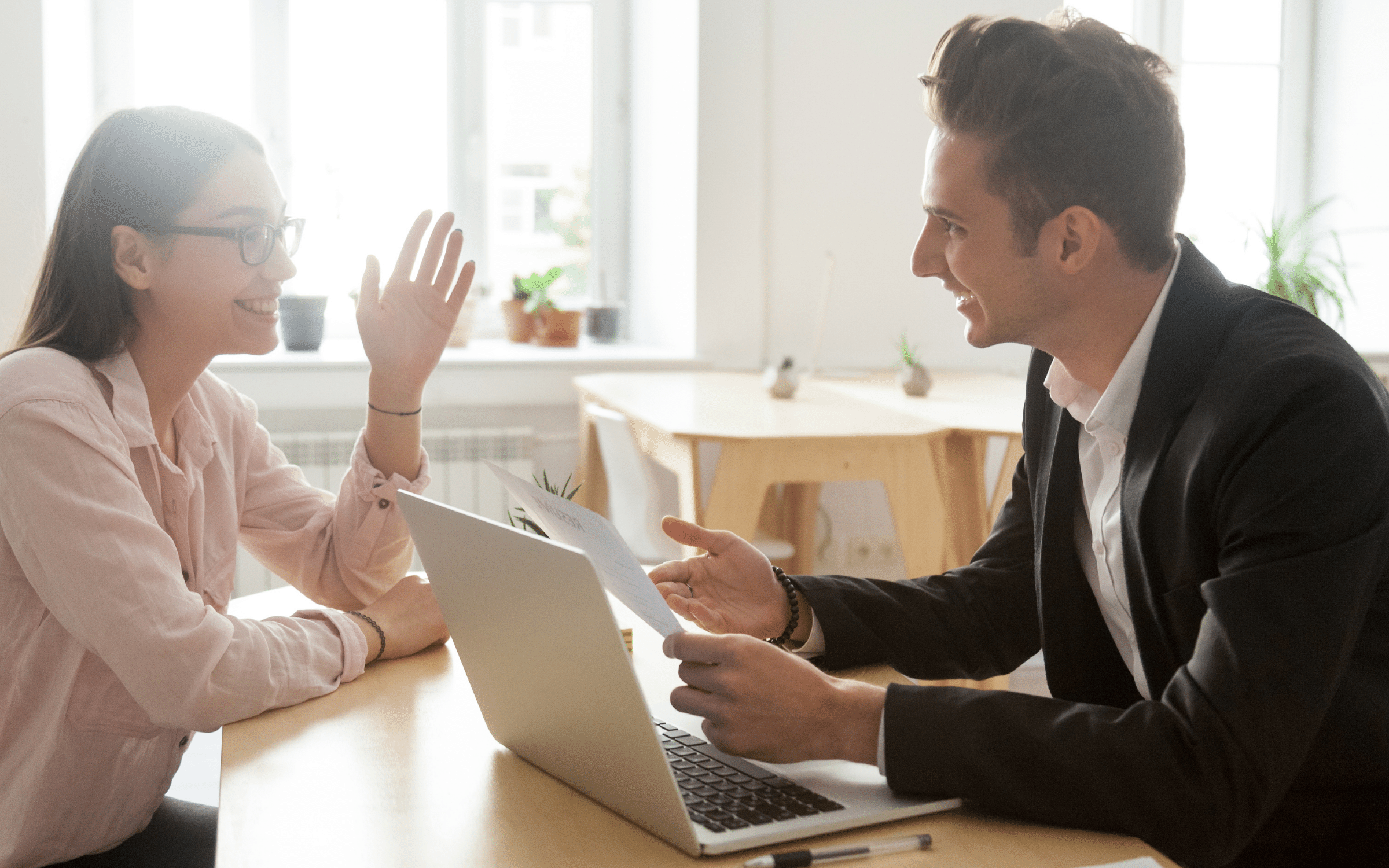 A professional man and woman engaging in a friendly discussion across a desk with a laptop, set in a bright office environment.