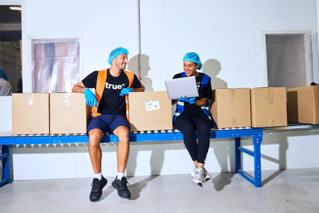 Two warehouse workers wearing safety vests, gloves, and hairnets sit on a conveyor belt among cardboard boxes, smiling and looking at a laptop.