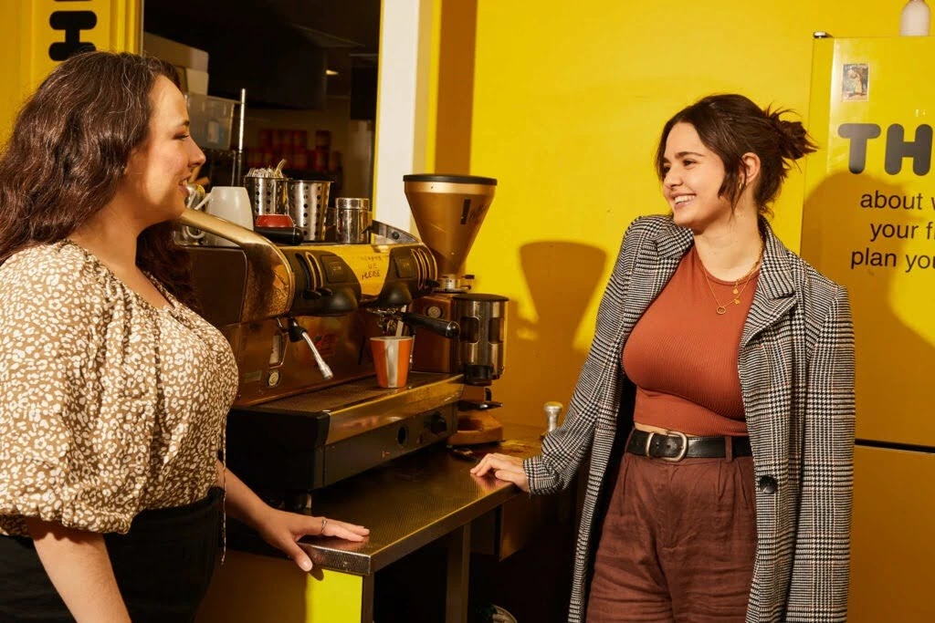 Two women happily chat in a vibrant yellow cafe. One leans on a counter near a coffee machine, while the other faces her, smiling warmly.