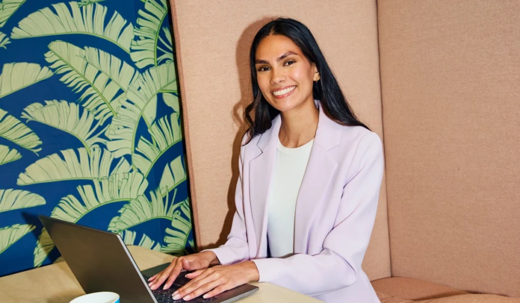 A smiling woman in a light purple blazer sits working on a laptop in a booth with tropical leaf-patterned wallpaper.