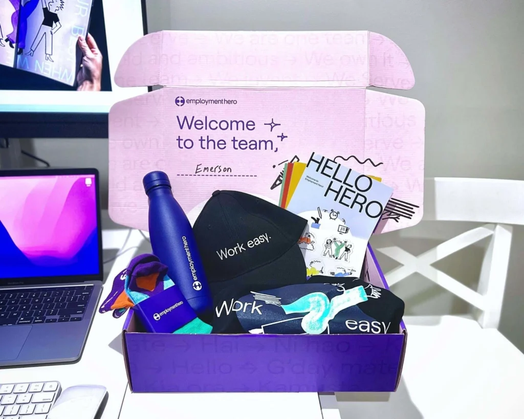 Gift box with "Welcome to the team, Emerson" text. Contains a blue water bottle, cap with "Work easy," socks, and a "Hello Hero" card. Bright, welcoming tone.