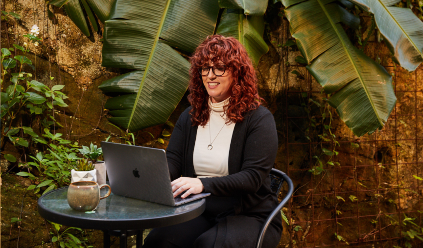 Woman sits at desk typing on her laptop in a casual garden environment