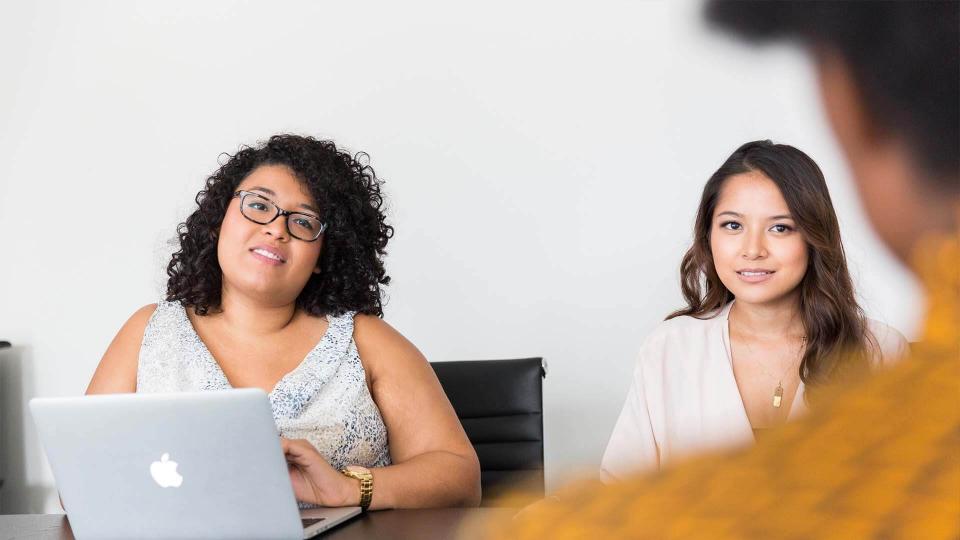 two women sitting behind the table with a laptop interviewing the third woman