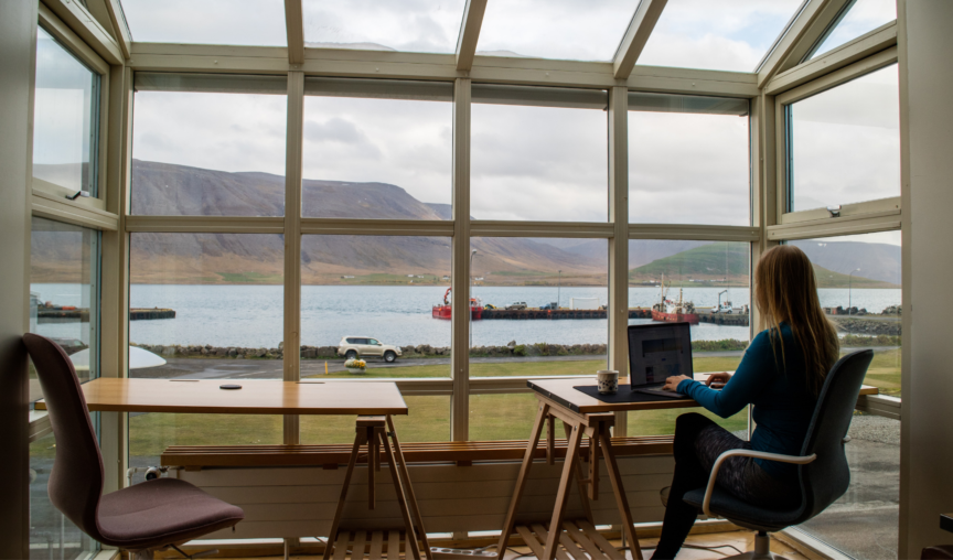 Woman sits at laptop in home office, overlooking a huge window that shows a lake and hills