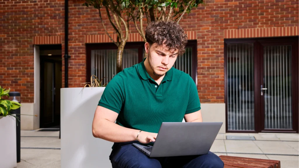 A young man with curly hair wearing a green polo shirt works attentively on a laptop computer outdoors in a brick courtyard setting.