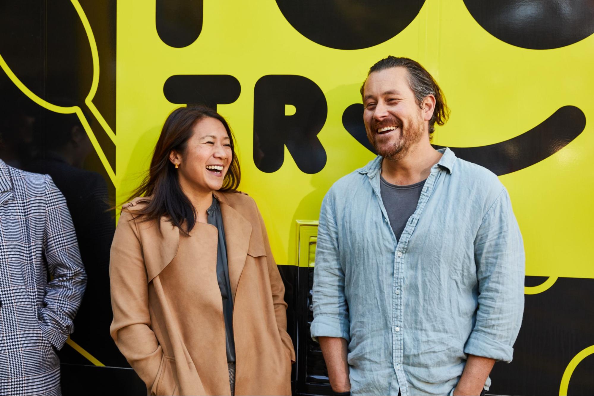 Two people, a woman in a tan coat and a man in a blue shirt, are laughing together in front of a large, bright yellow background with bold black text and a graphic.