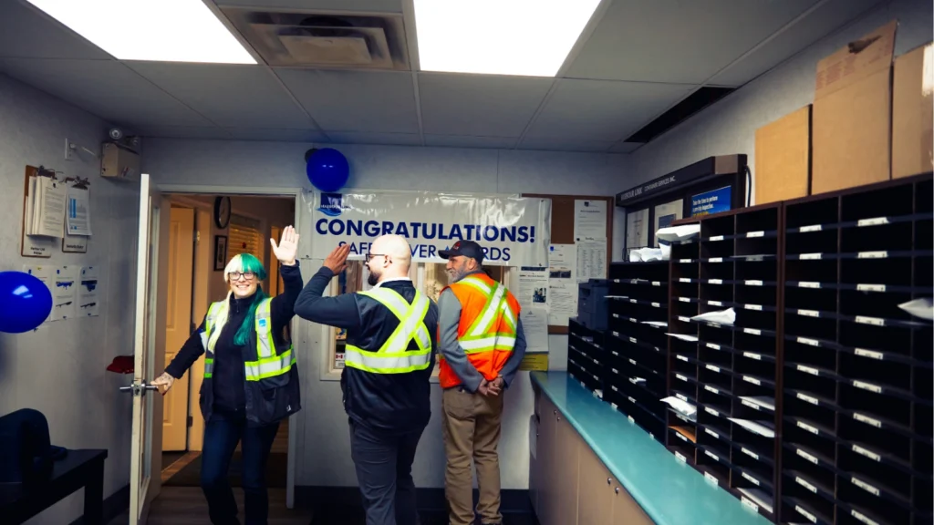 Industrial workers wearing high-visibility safety vests share a high five inside a office adorned with celebratory banners.
