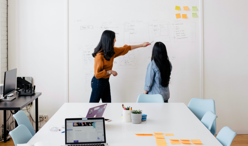 Two woman looking at a whiteboard, one pointing to findings on the board