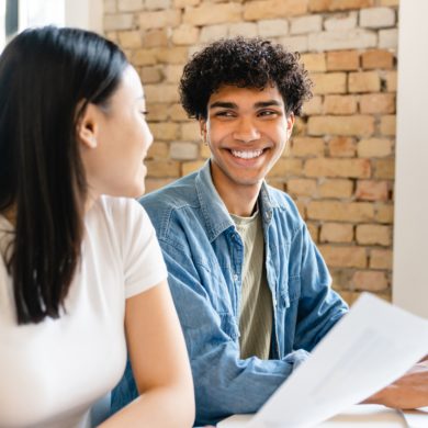 Two colleagues smiling and engaged in conversation while working together. One person is holding a piece of paper, and both are seated against a brick wall background, creating a collaborative and friendly work environment