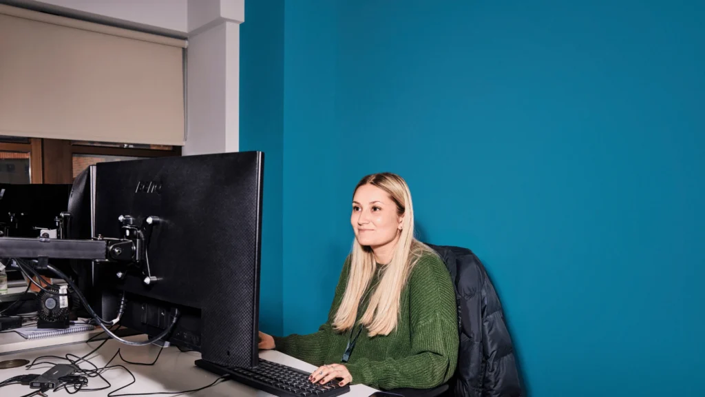 A young woman with long blonde hair sits at her desk in a modern office, smiling as she looks at a computer monitor against a vibrant teal wall.