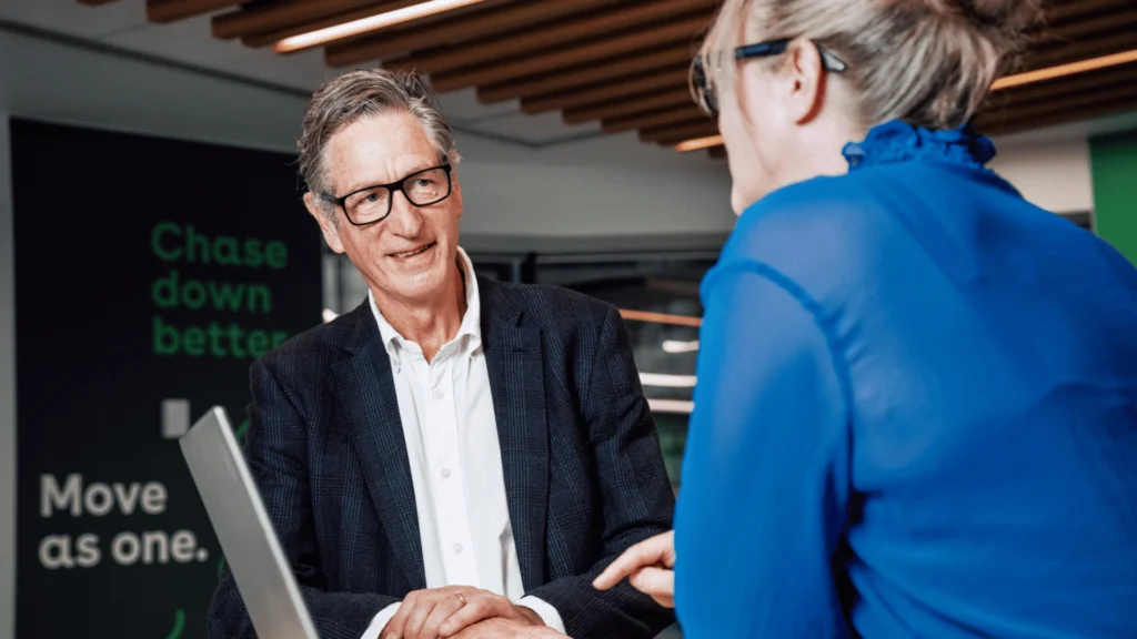 An older man wearing glasses and a suit jacket smiles while conversing with a woman in a blue blouse, sitting near a wall with motivational text.