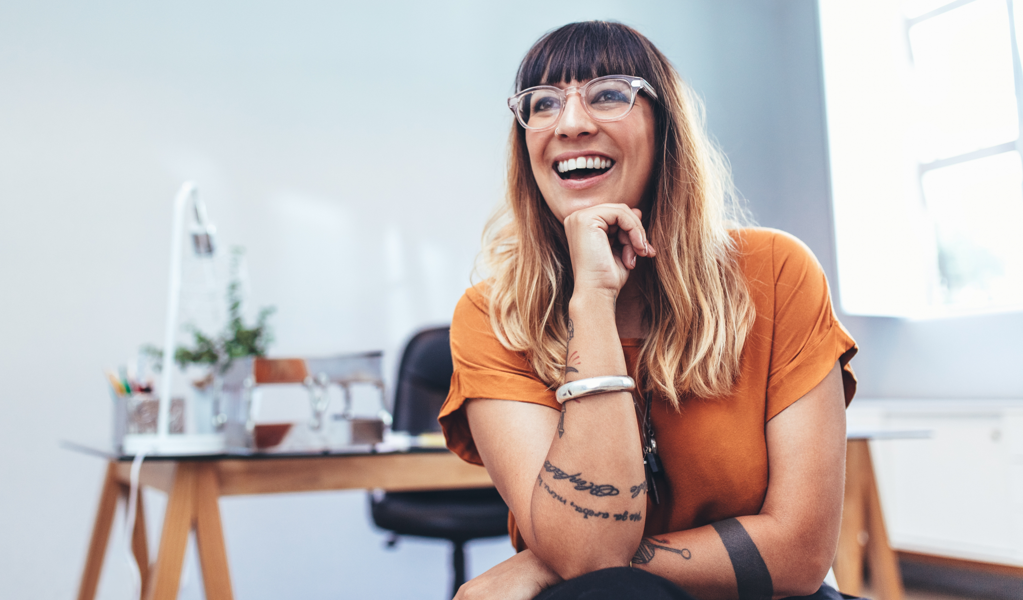 A smiling woman with glasses and tattoos sitting at a desk in a bright, modern office setting
