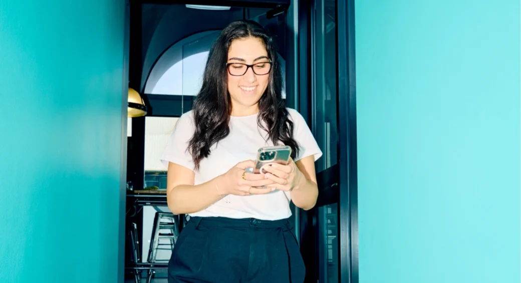 A woman with long dark hair and glasses stands in a bright hallway, smiling while looking at her phone. The teal walls create a vibrant, cheerful atmosphere.