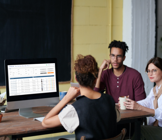 A group of professionals sitting around a table engaged in a meeting. A computer monitor on the table displays a data management or project tracking interface