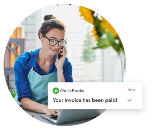 A photo of a woman wearing glasses and an apron, sitting at a desk while talking on the phone and using a laptop