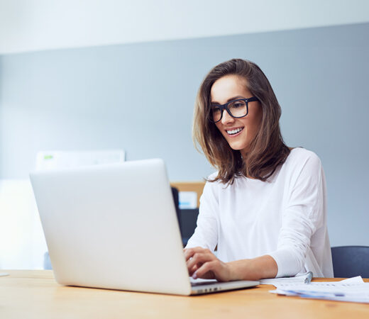 A photo of a woman wearing glasses, smiling as she works on a laptop in a well-lit office, conveying a sense of focus and satisfaction