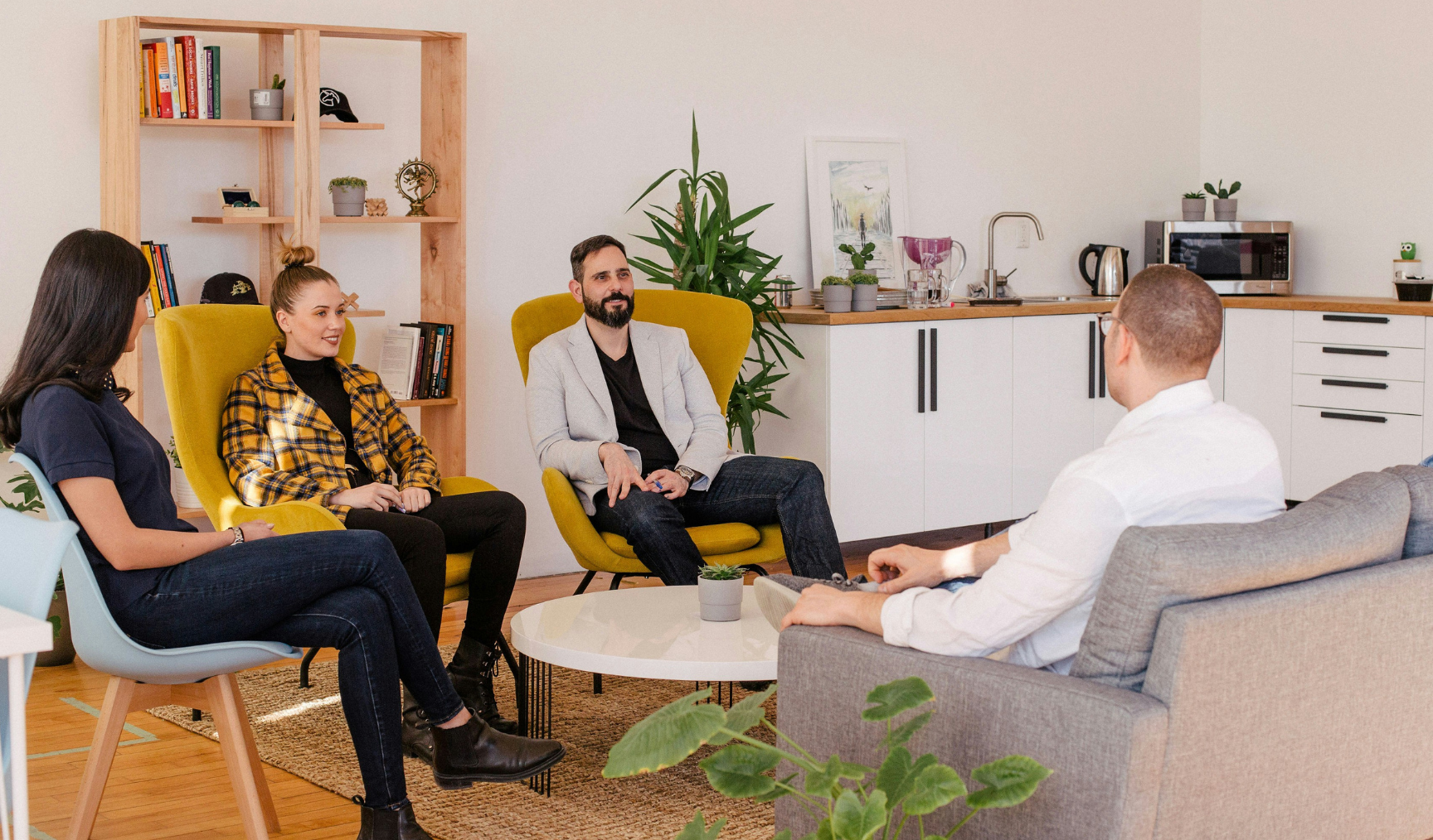 A group of people sit in a casual meeting room, on comfy chairs around a coffee table