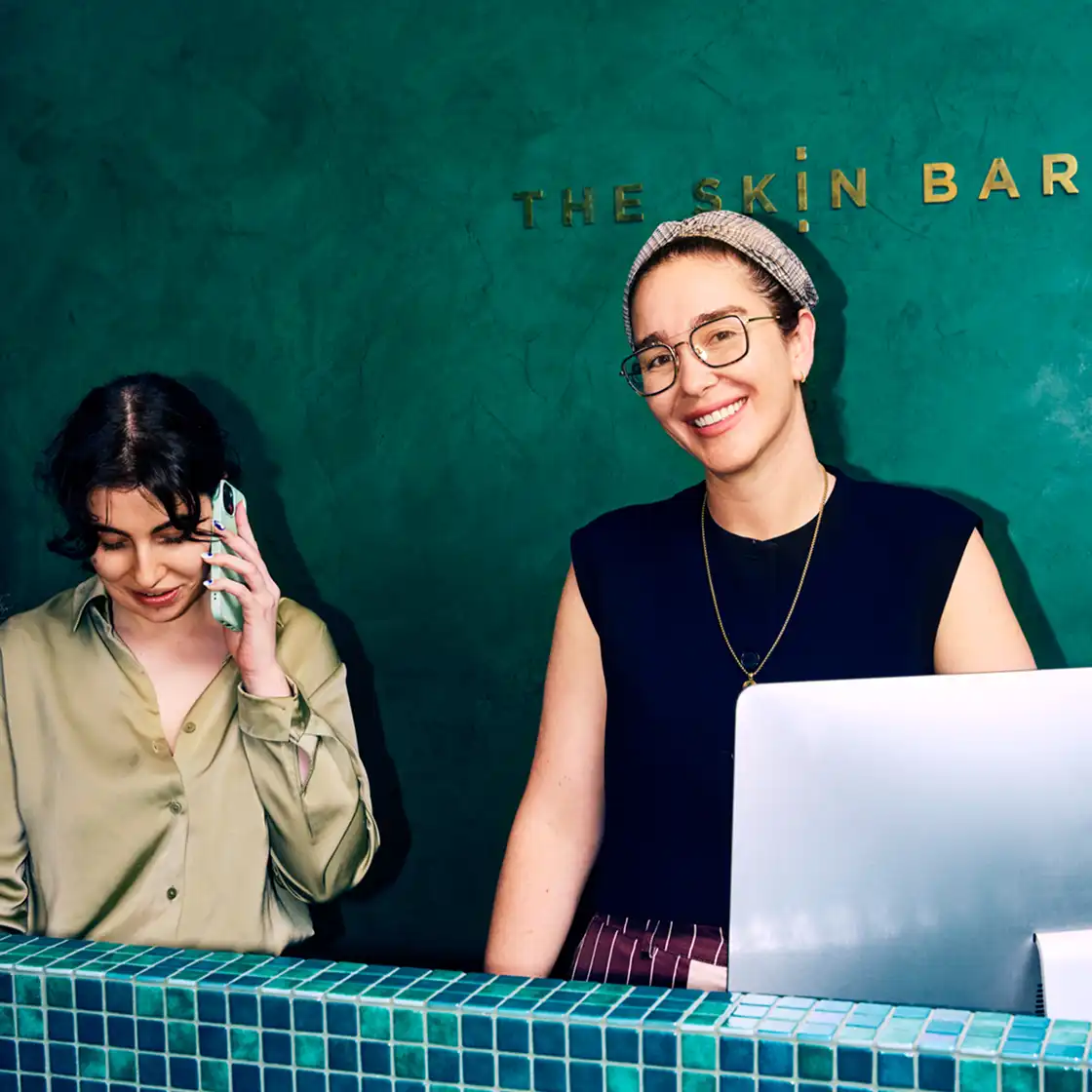 Two women at a green-tiled reception desk. One is on the phone, wearing a light blouse. The other smiles behind a computer, with "The Skin Bar" sign.