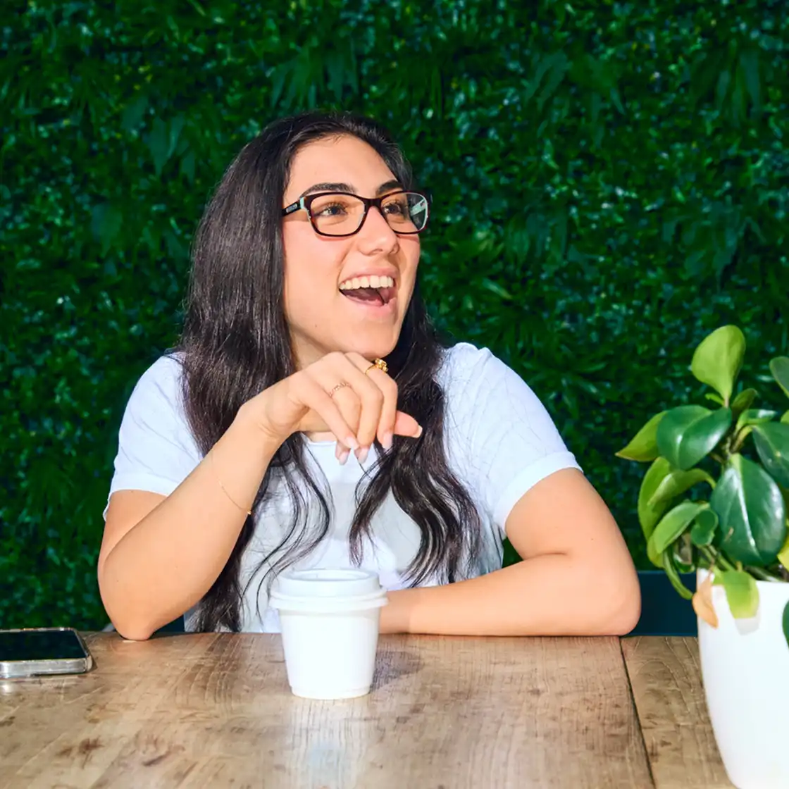 Woman with glasses laughing joyfully, sitting at a wooden table with a small white coffee cup. Green leafy background and a potted plant beside her.