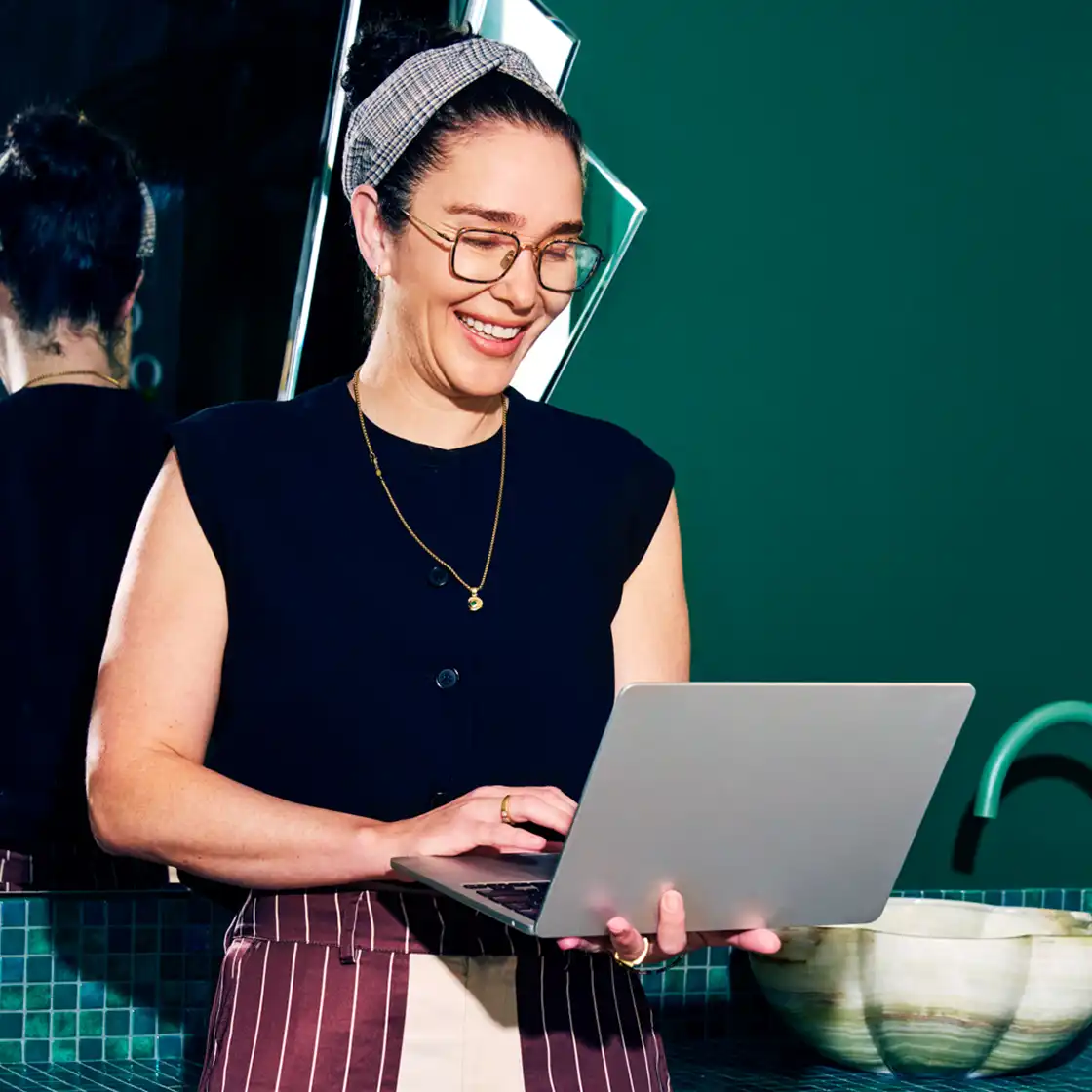 A smiling person with glasses and a headband stands holding a laptop. They wear a dark top and striped apron in a modern green-tiled room.