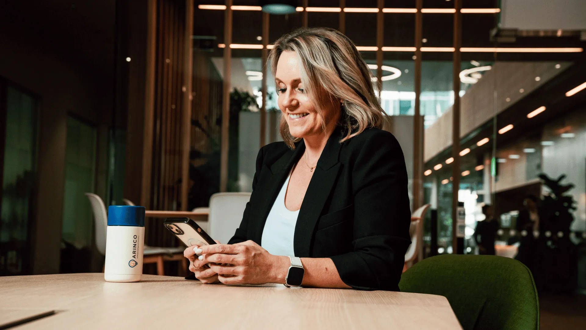 A smiling female professional in a black blazer checking her smartphone at a modern office desk, representing an HR manager staying organised ahead of the End of Financial Year.