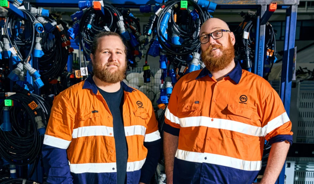 Two industrial workers wearing high-visibility orange uniforms standing in front of organized cables in a workshop or warehouse.

