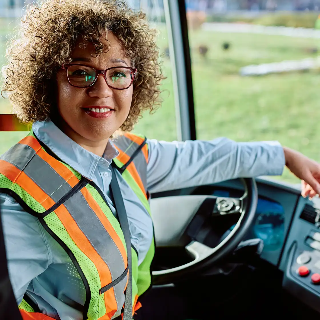 A smiling bus driver with curly hair and glasses, wearing a reflective safety vest, sits confidently at the wheel, evoking a sense of professionalism.