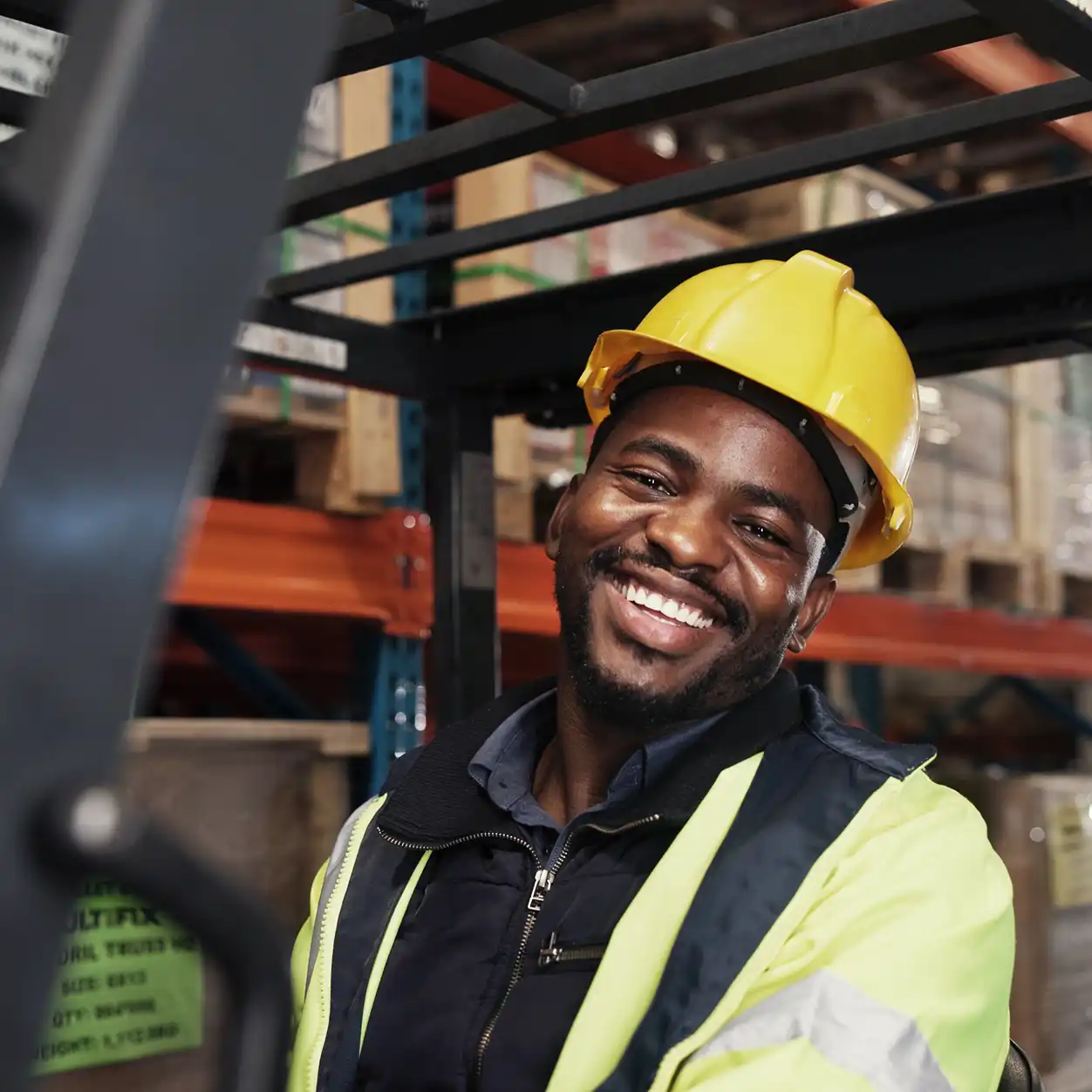 Smiling worker in a yellow hard hat and reflective jacket sits in a warehouse. Background shows shelves stacked with boxes, conveying a positive, industrious vibe.