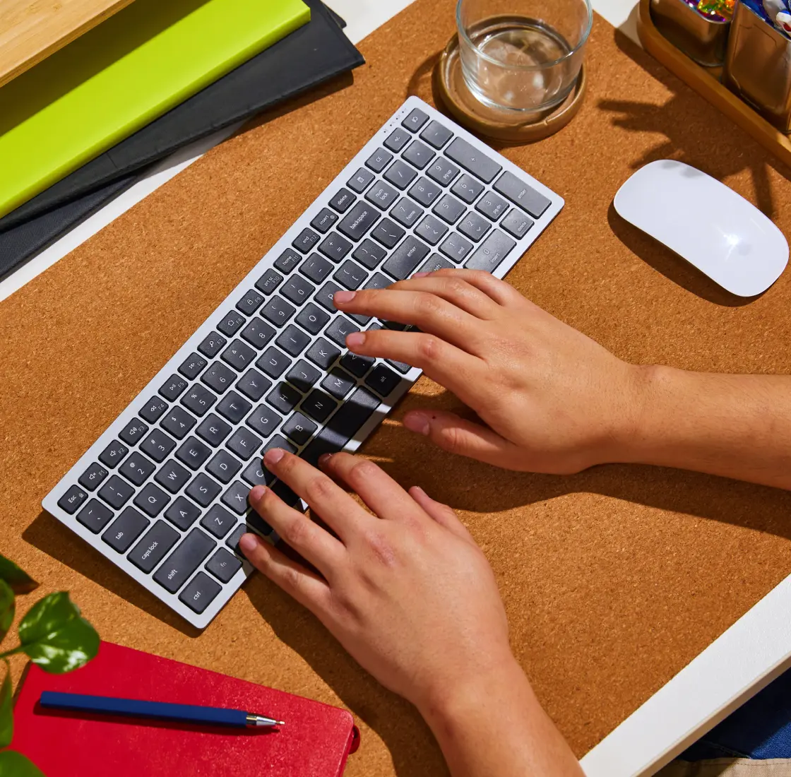 Close-up of hands typing on a wireless keyboard at a tidy desk setup with a mouse, notebooks, and pen.