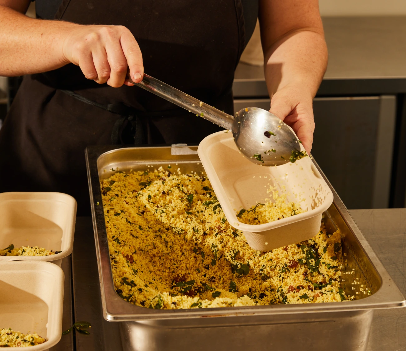 Close-up of a person in an apron serving couscous into a takeaway container in a kitchen.