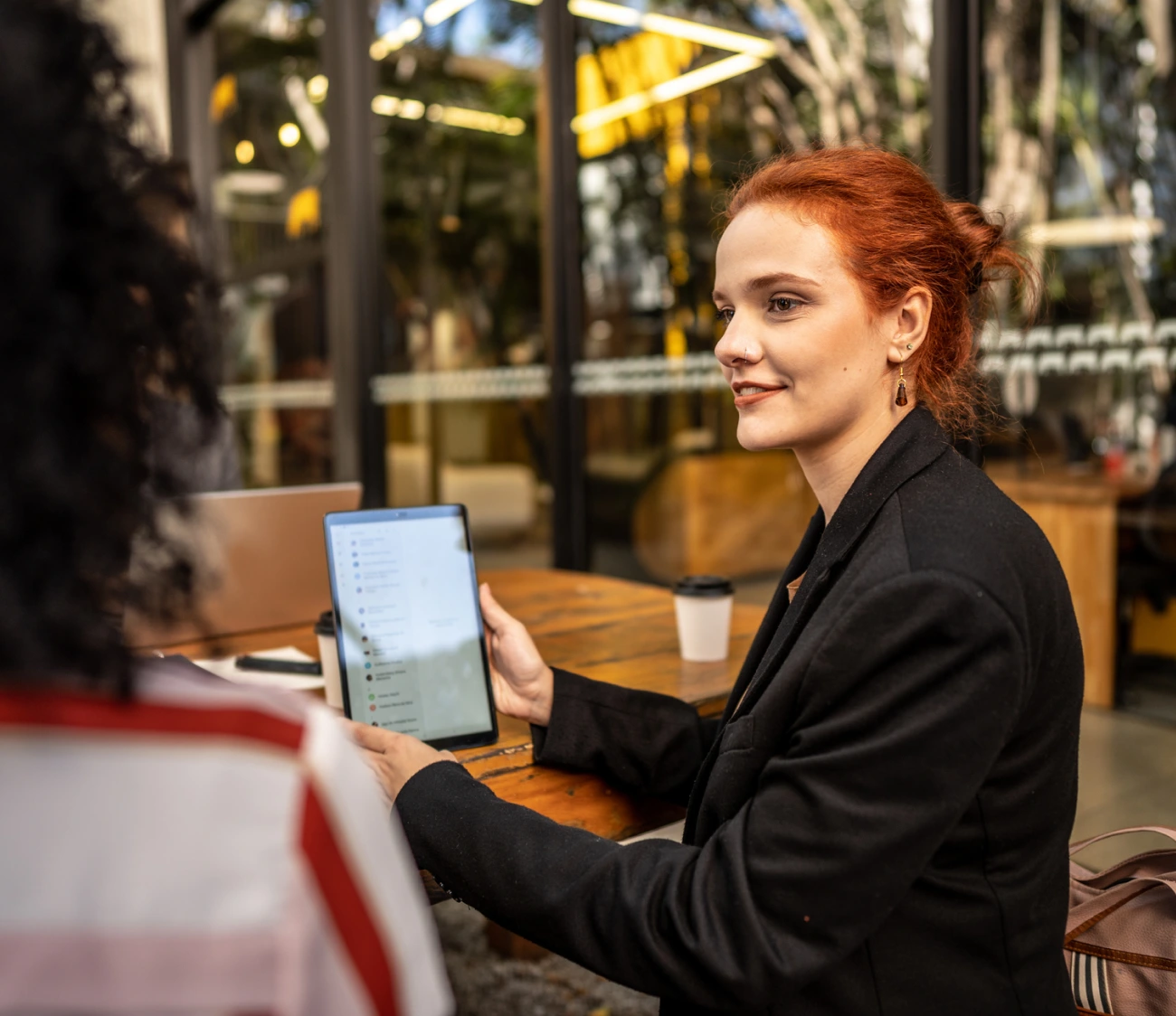 Woman with red hair holding a tablet and talking to someone across a table in an outdoor setting.