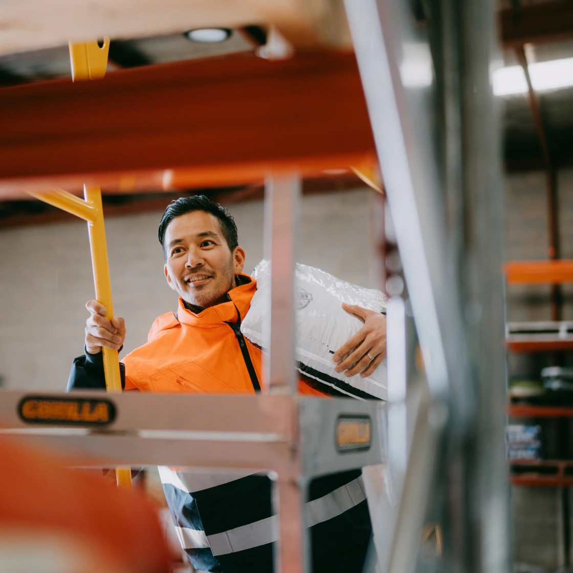 A smiling warehouse worker in a reflective orange and black jacket carries a large white bag on his shoulder while holding a yellow ladder in an industrial setting.