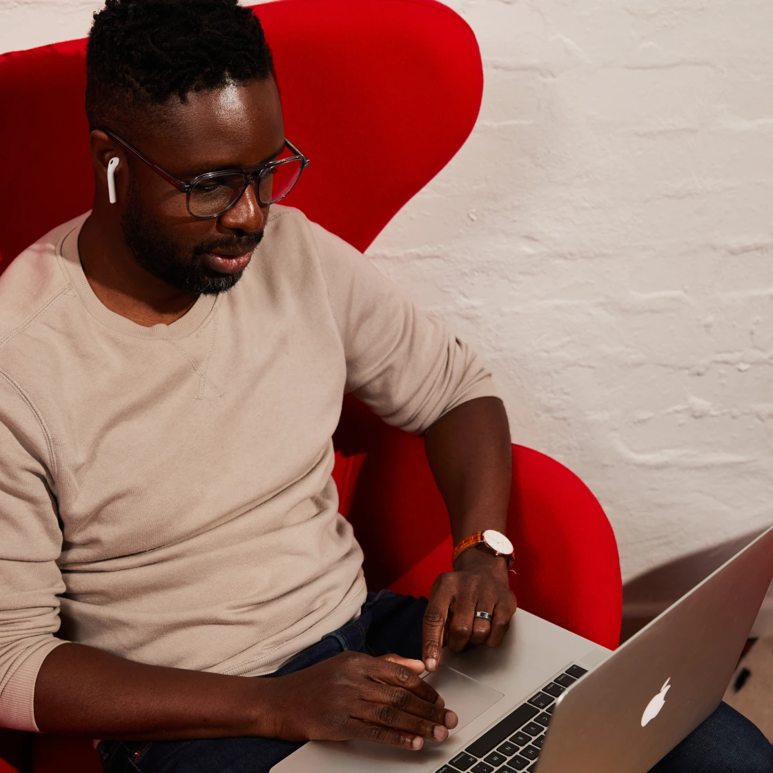 A man wearing glasses, wireless earbuds, and a beige sweater sits in a bold red chair while using a silver MacBook laptop.