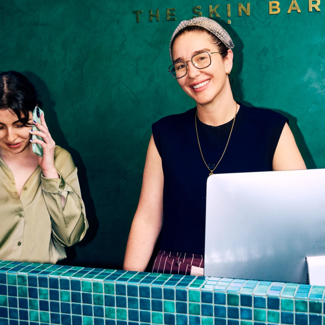 Two women at a tiled reception counter, one smiling at the camera and the other on the phone, with "The Skin Bar" signage behind them.