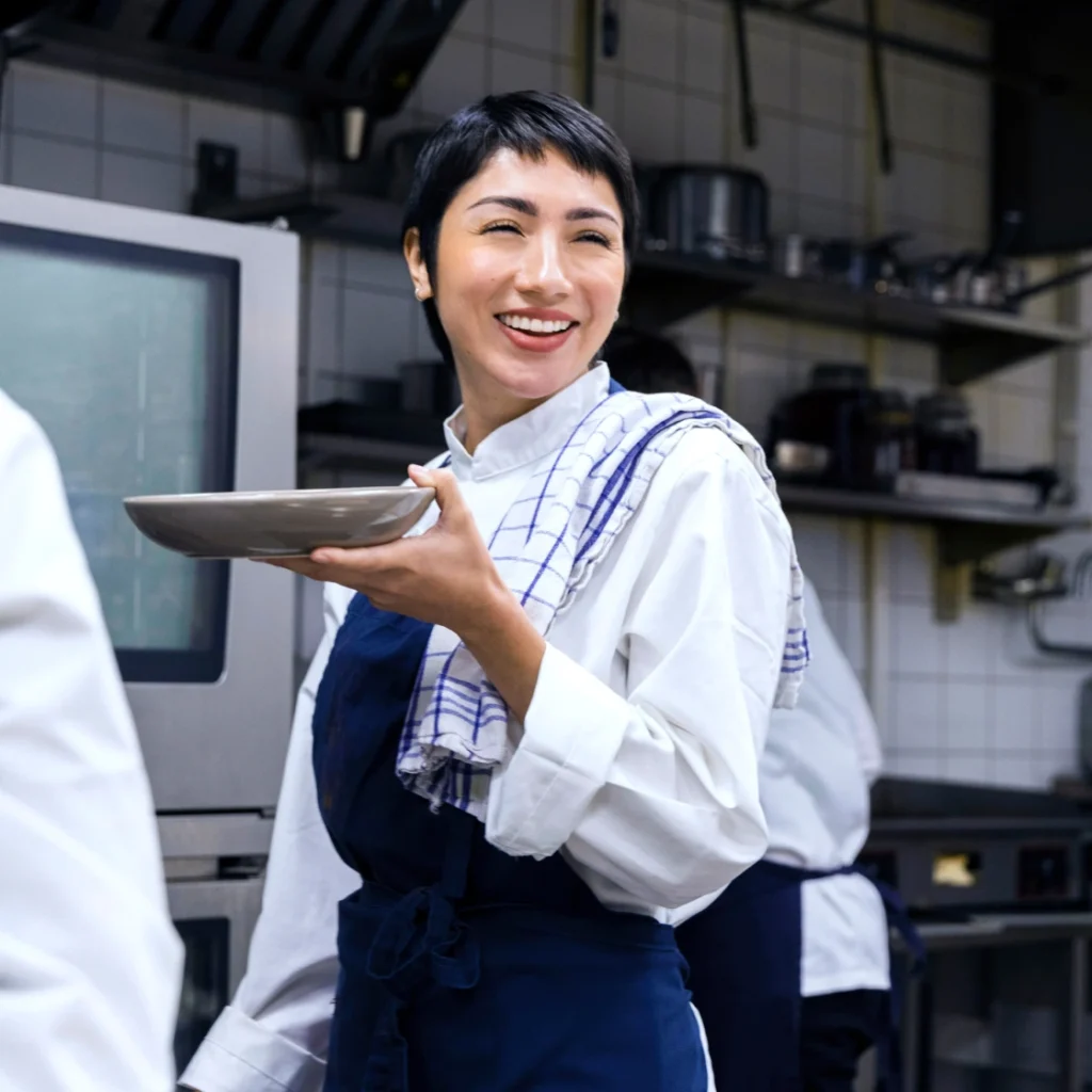 A cheerful chef in uniform holding a plate with a towel over her shoulder, standing in a commercial kitchen.