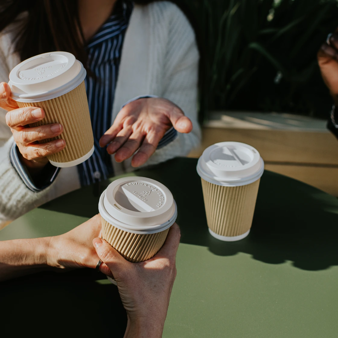 Three people seated around a green table holding takeaway coffee cups, engaged in conversation.