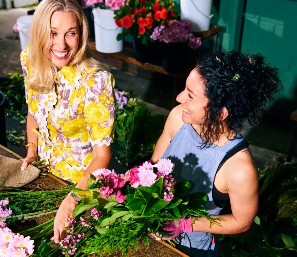 Two women smile while arranging vibrant flowers at a florist workspace, surrounded by flower buckets and greenery.