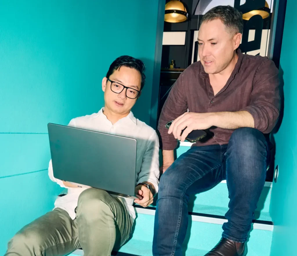 Two men sit on stairs looking at a laptop screen, seemingly discussing or reviewing content.