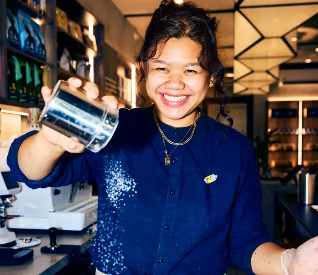 A smiling barista shakes a metal container, possibly sprinkling ingredients, in a well-lit, modern cafe.