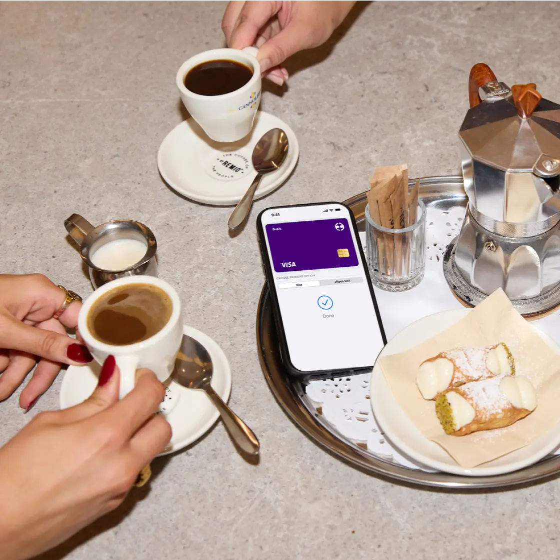 A table scene with two people having coffee and desserts; a smartphone on the tray displays the Visa card in a mobile wallet app.