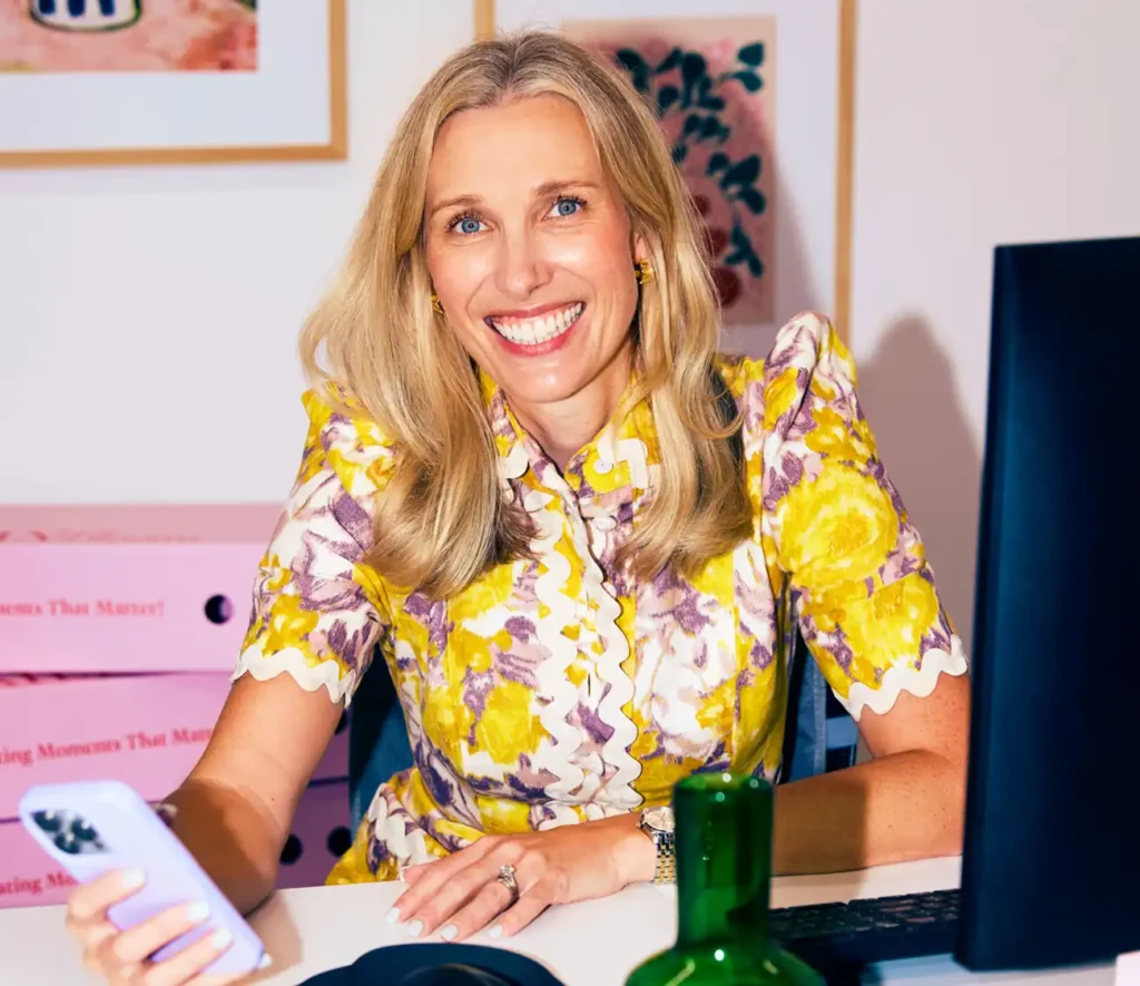 A woman in a floral dress sits at a desk, holding a cell phone and looking thoughtfully at her workspace.