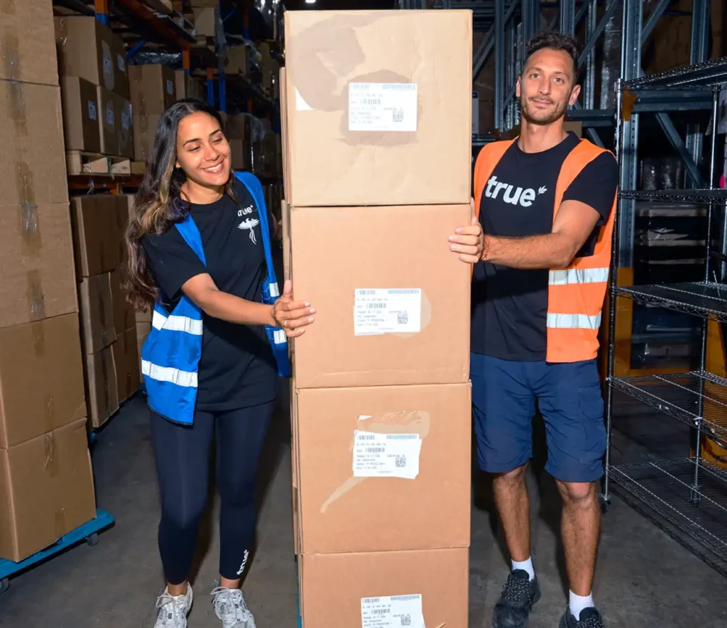 Two people stand beside a large box in a warehouse, surrounded by shelves and industrial equipment.