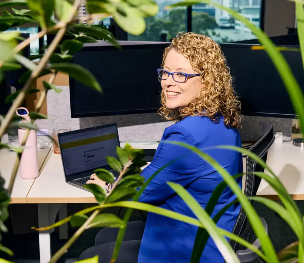 A woman sitting at a computer with green plants in the background, focused on her work.