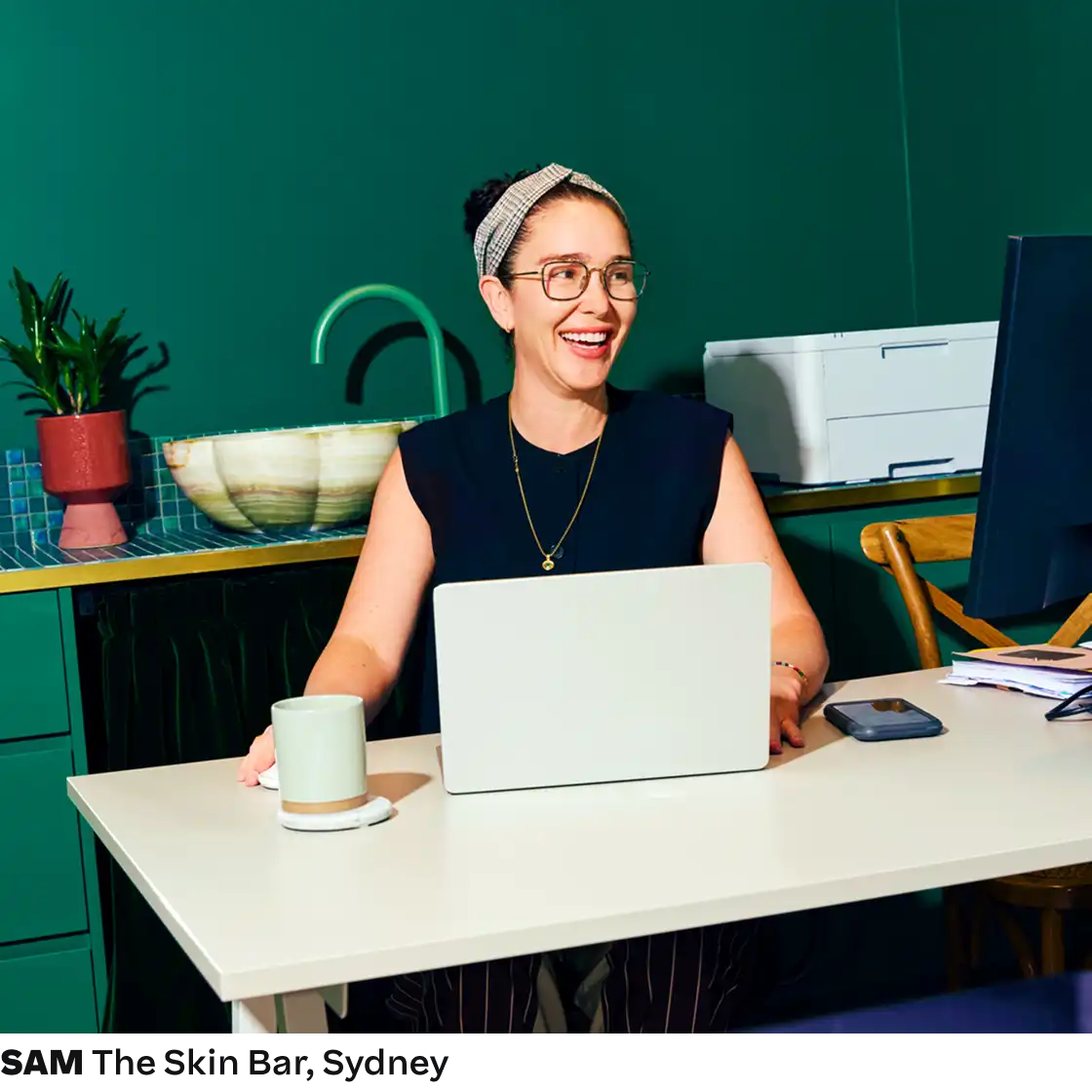 A woman with glasses and a headband smiles while sitting at a desk with a laptop and coffee. The room is colorful with a green wall and plants.
