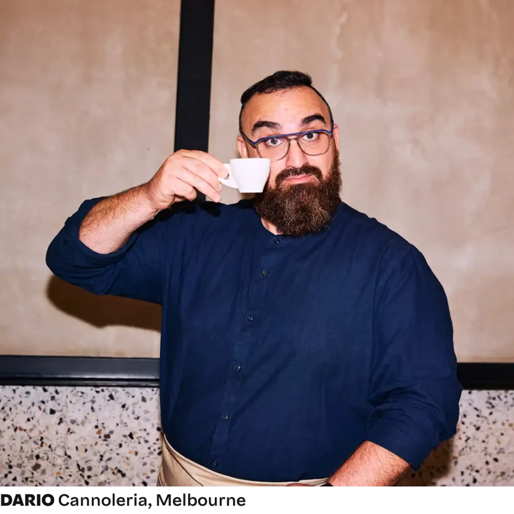 A man with a beard and glasses humorously holding a small white cup up to his eye. He's wearing a blue shirt, standing against a speckled wall.