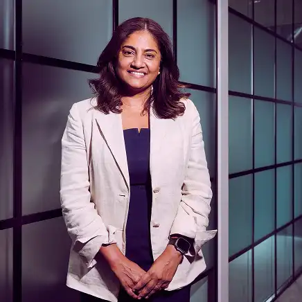 A woman in a light blazer and dark top smiles confidently, standing in front of frosted glass panel walls. The setting is modern and professional.