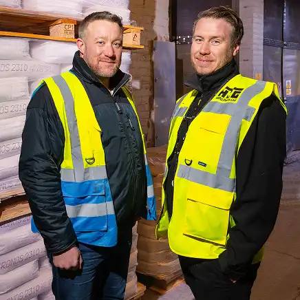 Two men in yellow safety vests stand in a warehouse with stacks of bags and wooden pallets. They appear confident and professional.
